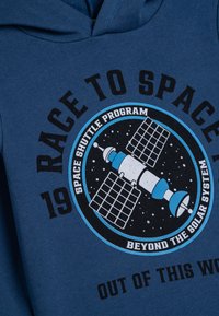 Felpa blu con cappuccio caratterizzata da una grafica circolare di un satellite, con testo in bianco e nero che recita "CORSA PER LO SPAZIO" e "FUORI DA QUESTO MONDO."