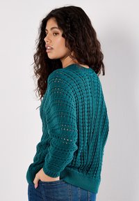 Maglione lavorato a maglia color teal con motivo testurizzato e trame aperte, dotato di polsini e orlo a costine. Modello mostrato di lato.