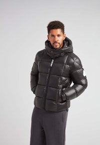 Giacca a vento nera con collo alto e cappuccio regolabile, caratterizzata da un design trapuntato, chiusura frontale con zip e dettaglio del logo sul petto.