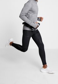Maglia atletica grigia a maniche lunghe e leggings neri aderenti con tasche laterali, abbinati a scarpe da corsa bianche, indossati da una persona in movimento.