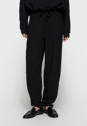 Person iført løse sorte bukser med snøre i taljen og sorte læderloafers mod en ensartet lys baggrund.