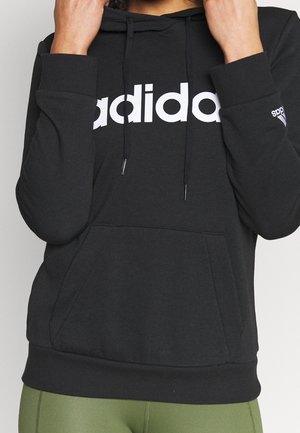 Personne portant un sweat à capuche Adidas noir avec un logo blanc, les mains ajustant les cordons de la capuche, associé à des leggings verts pour l'entraînement.