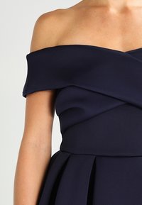 Marinblå off-shoulder klänning med figurnära bodice, inlindad design och fl ödande kjol. Slät tyg utan synliga mönster eller fästen.