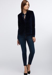 Navy velvet blazer with narrow lapels, front pockets, and a fitted silhouette, paired with dark blue skinny jeans and black high heels.
