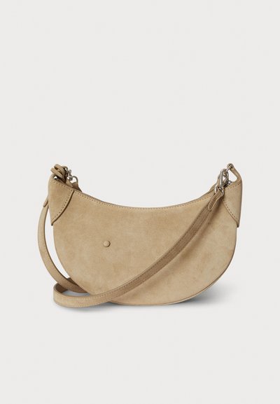 Beżowa zamszowa torebka w kształcie półksiężyca, z zamkiem błyskawicznym i regulowanym paskiem. Gładka faktura i minimalistyczny design z niewielkim detalem w postaci kropki.
