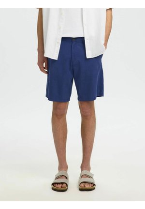 Homme portant un short bleu, une chemise blanche et des sandales beiges, debout devant un fond clair uni, les mains dans les poches.