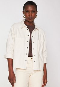 Camicia di denim beige chiaro con design a collare, bottoni a scatto e tasche sul petto. Indossata su una maglietta marrone scuro e abbinata a pantaloni color crema.