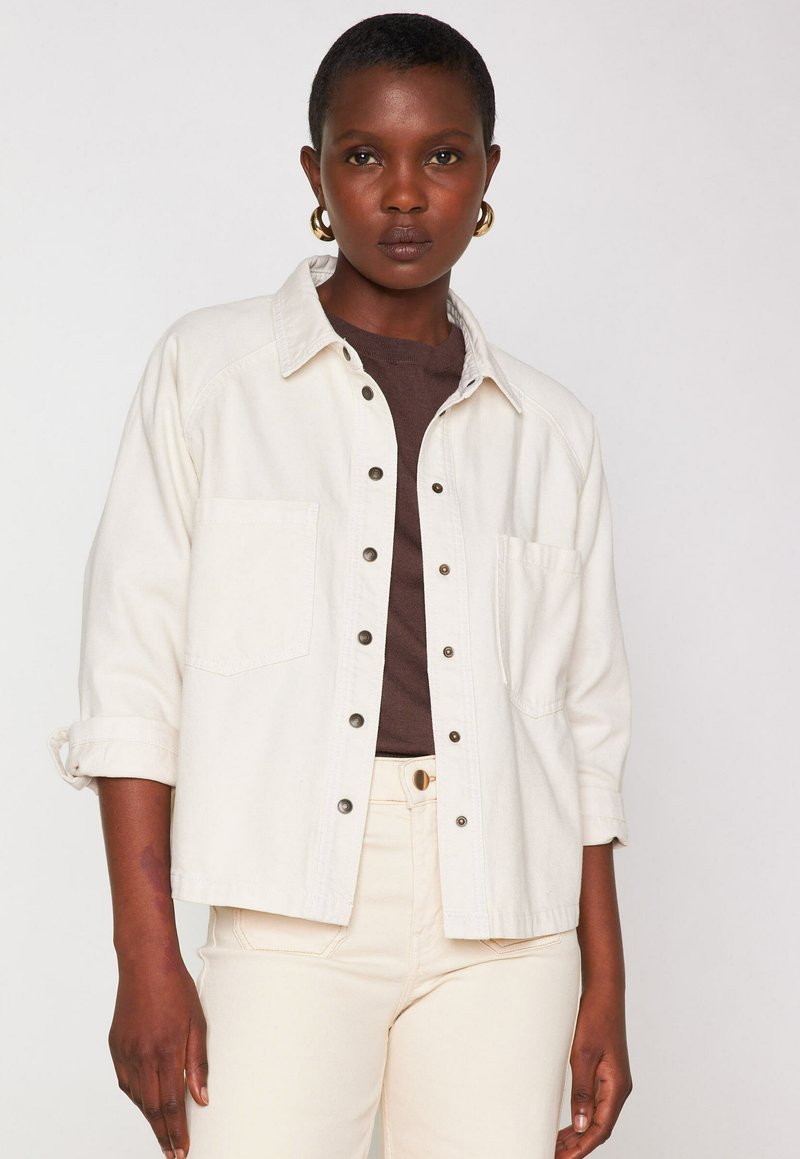 Camicia di denim beige chiaro con design a collare, bottoni a scatto e tasche sul petto. Indossata su una maglietta marrone scuro e abbinata a pantaloni color crema.