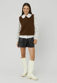 Femme souriante portant un pull sans manches marron par-dessus un chemisier floral, un short en cuir noir et des bottes blanches jusqu'au genou, se tenant devant un fond uni.