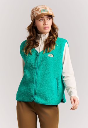 Vrouw draagt een turquoise fleecevest over een crèmekleurige longsleeve, bruine broek en een beige pet met patroon, licht naar rechts gericht.