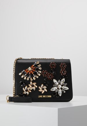 Sac bandoulière noir Moschino avec ornements floraux en strass multicolores et bandoulière chaîne dorée sur un socle blanc.