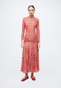 Robe à manches longues aux tons roses et rouges, présentant un motif texturé, un corsage ajusté et des détails drapés à la taille. Chaussures plates rouges.