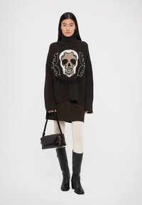 Brauner oversized Pullover mit Totenkopfdesign, dunkler Rock, weiße Strumpfhosen und schwarze kniehohe Stiefel, ergänzt durch eine strukturierte braune Handtasche.