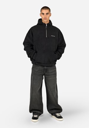 Črna flisanje kapuca z kenguru žepom, srebrnim zadrgo in logotipom, v kombinaciji s temnimi širokimi hlačami in črnimi teniskami.