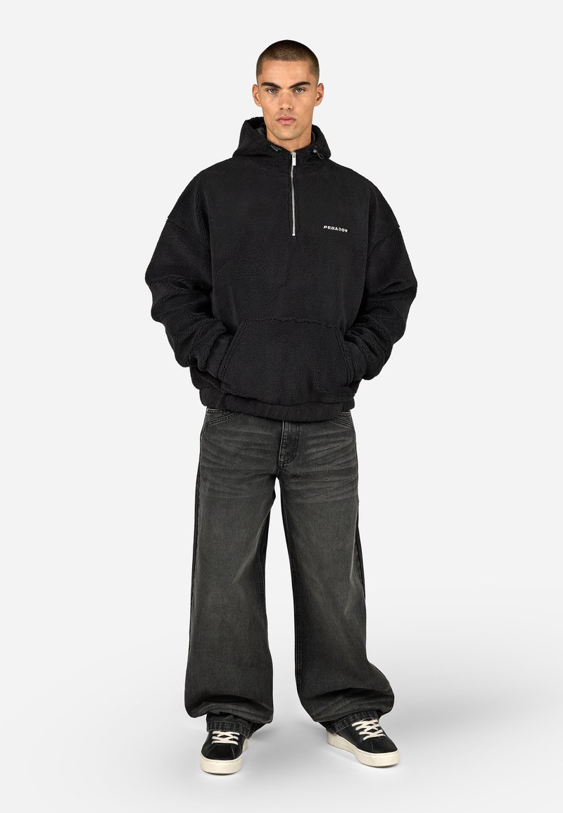 Musta fleece huppari, jossa on kengurupussi, hopeinen vetskari ja logotiedot, yhdistettynä tummiin leveisiin farkkuihin ja mustiin lenkkareihin.