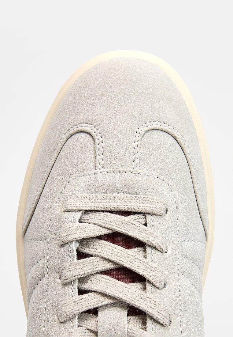 Jasnoszare zamszowe trampki z okrągłym noskiem, sześcioma dziurkami na sznurowadła, teksturowaną powierzchnią i kontrastującymi bordowymi sznurówkami. Guma widoczna na spodzie.