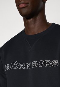 Μαύρη μπλούζα με στρογγυλή λαιμόκοψη, με λευκό κείμενο "BJÖRN BORG" σε έντονη, σύγχρονη γραμματοσειρά στο στήθος. Το κείμενο περιλαμβάνει τον διασκορπισμένο τόνο.