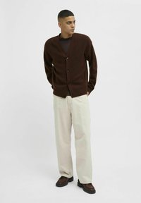 Brown cardigan made from a textured material, featuring a V-neck, five buttons down the front, and ribbed cuffs, paired with cream pants.