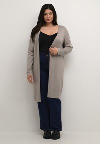 Cardigan lungo beige con maniche lunghe, apertura frontale e polsini a coste, indossato sopra un top fitted nero e jeans a gamba larga blu scuro.