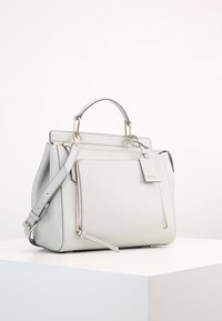 Jasnoszara, strukturalna torebka skórzana z górnym uchwytem, regulowanym paskiem na ramię, przednią kieszenią na zamek błyskawiczny oraz zwisającą etykietą na białej powierzchni.