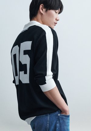 Mladá osoba v černém svetru s bílými pruhy a číslem "05" na zádech, ruce v kapsách, dívá se dolů na jednoduchém pozadí.