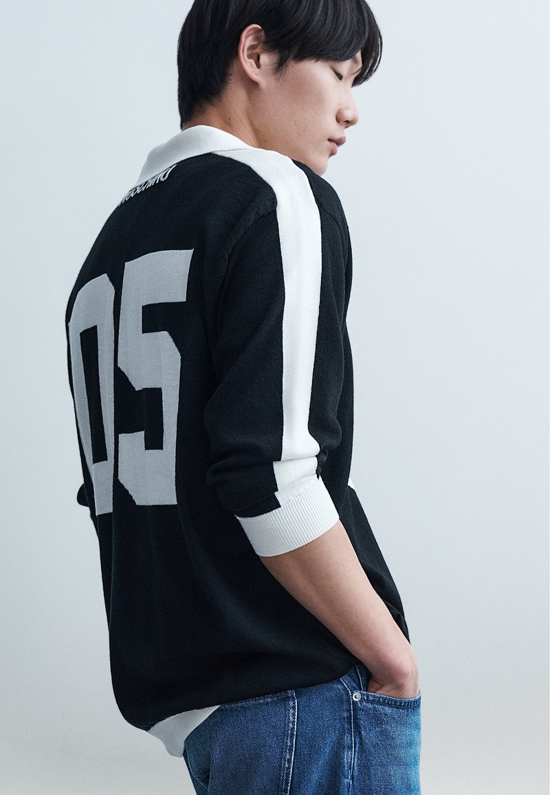 Νέος άνθρωπος που φοράει μαύρο πουλόβερ με λευκές ρίγες και το "05" στην πλάτη, με τα χέρια στις τσέπες, κοιτάζοντας προς τα κάτω σε απλό φόντο.