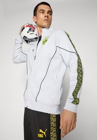 Homme portant une veste de football BVB blanche et un pantalon noir Puma, tenant un ballon de football Puma sur son épaule contre un fond uni.