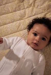 Cardigan blanc avec un motif en torsades porté par un bébé, allongé sur une couverture jaune à motifs floraux. Les cheveux du bébé sont bouclés et foncés.