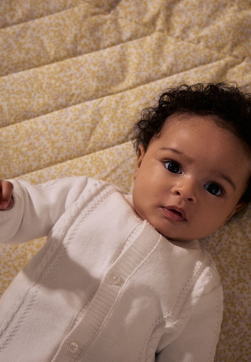 Cardigan blanc avec un motif en torsades porté par un bébé, allongé sur une couverture jaune à motifs floraux. Les cheveux du bébé sont bouclés et foncés.