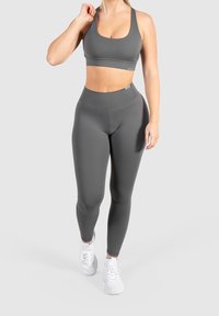 Mujer vestida con un sujetador deportivo gris y leggings de cintura alta, caminando hacia adelante con zapatillas blancas sobre un fondo liso.