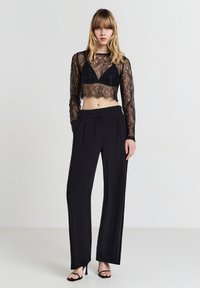Frau steht da und trägt ein schwarzes Spitzen-Crop-Top über einem Bralette, hochgeschnittene schwarze weit geschnittene Hosen und schwarze High Heels.
