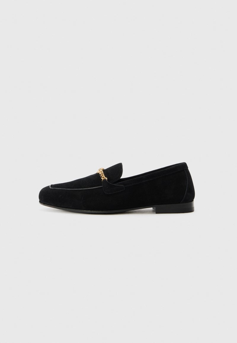 Mocassins en daim noir avec un talon plat, présentant un accent décoratif en chaîne dorée à l'avant et un design à bout arrondi.