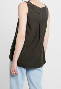 Haut sans manches vert olive foncé avec un col rond, un détail dos en forme d'œillet, et un drapé léger ; associé à un pantalon large bleu clair.