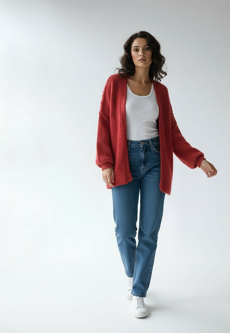 Czerwony, pluszowy kardigan na białym topie na ramiączkach, w połączeniu z niebieskimi dżinsami i białymi trampkami. Kardigan ma opuszczone ramiona i luźny krój.