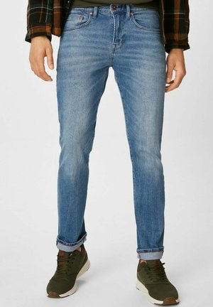 Mann trägt hellblaue, eng anliegende Jeans mit aufgekrempelten Säumen, olivgrüne Sneakers und eine kariertes Jacke, steht mit entspannten Händen an den Seiten.