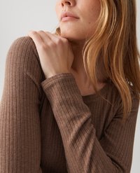 Woman with light brown hair wearing a ribbed, long-sleeve brown top, holding her shoulder with her left hand against a plain background.