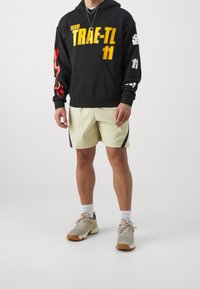 Sweat à capuche noir avec des motifs jaunes et rouges, assorti à un short beige clair avec des accents latéraux noirs et des chaussures de sport grises.
