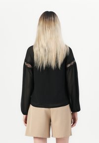 Persona con lunghi capelli biondi che indossa una blusa nera trasparente a maniche lunghe con dettagli in pizzo e pantaloni beige, in piedi contro uno sfondo bianco.