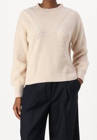 Pull en tricot beige avec un motif en câble, col rond, poignets et ourlet côtelés, adopte une coupe décontractée. Assorti à un pantalon foncé.