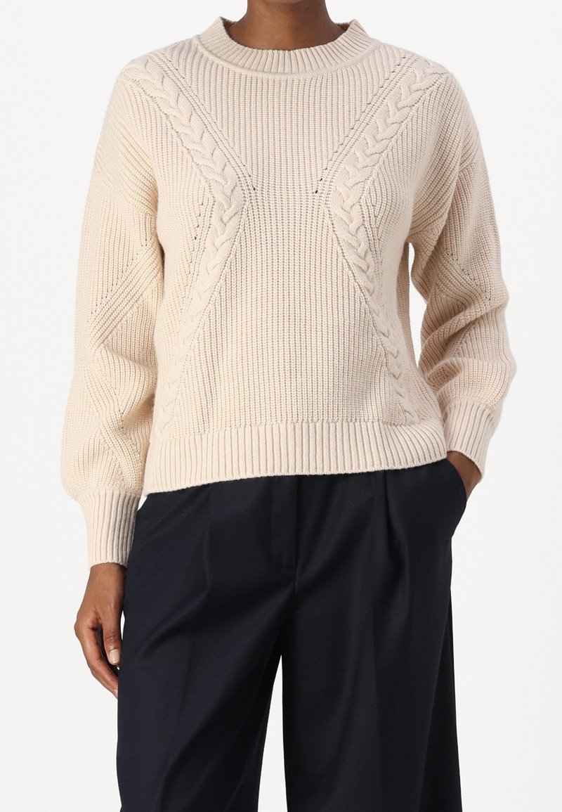 Pull en tricot beige avec un motif en câble, col rond, poignets et ourlet côtelés, adopte une coupe décontractée. Assorti à un pantalon foncé.