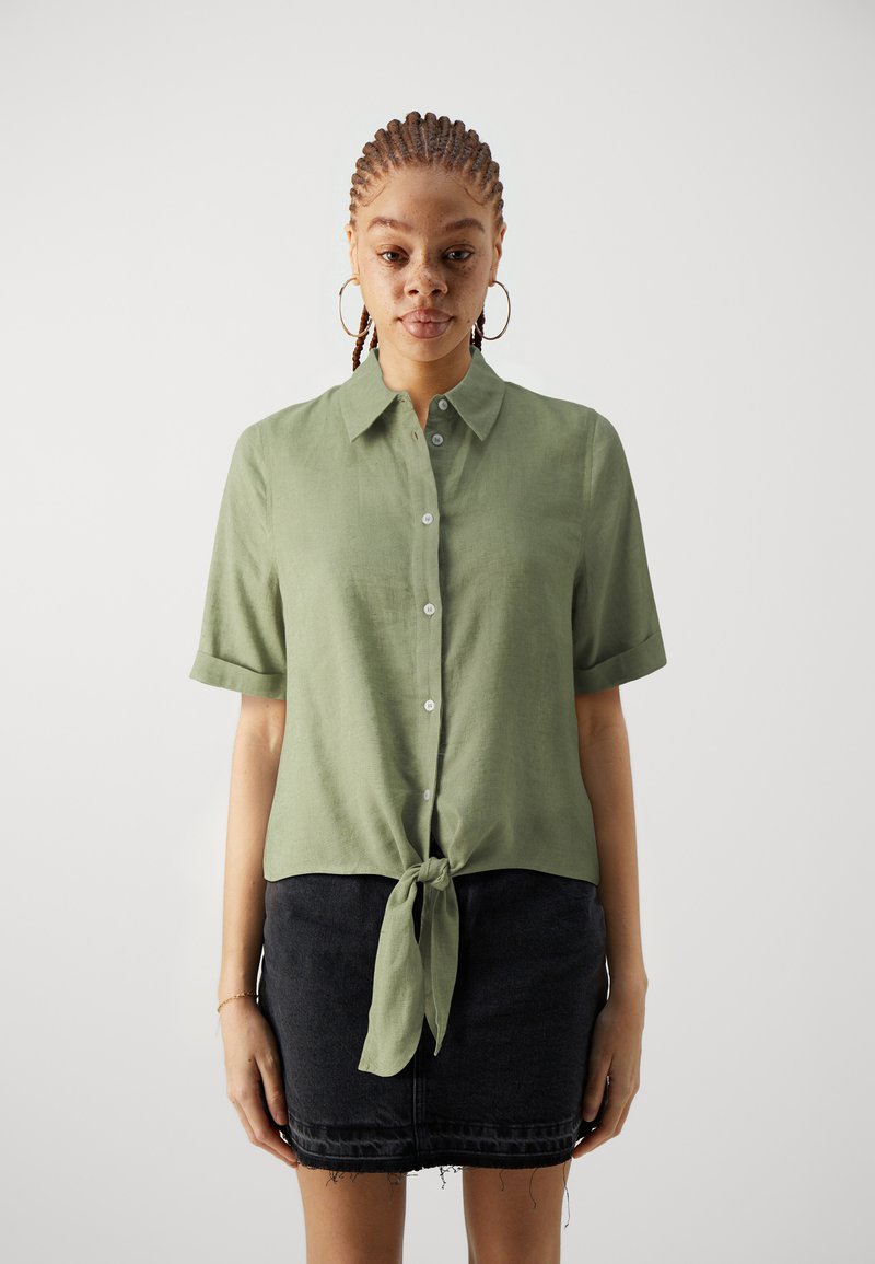 Korte mouwen groene linnen shirt met knoopsluiting aan de voorkant en een gestrikte taille; gecombineerd met een zwarte denimsrok met rafelige zoom.