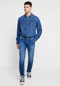 Denim shirt met twee borstzakken, knoopsluiting aan de voorkant, klassieke kraag, gecombineerd met slim-fit denim jeans, licht vervaagd blauw, omgeslagen bij de enkels, zwarte sneakers.