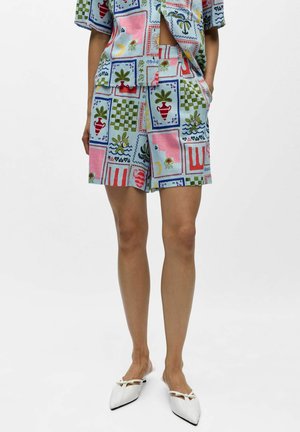 Osoba oblečená v pestrofarebných krátkych šortkách s potlačou a zladenou krátkym rukávom, doplnená bielymi plochými topánkami so špicatou špičkou a malými mašličkami.