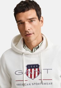 Άνδρας με κοντά σκούρα μαλλιά που φοράει λευκό φούτερ με κουκούλα με σήμα ασπίδας σε κόκκινο, άσπρο και μπλε χρώμα και το κείμενο "GANT 1949 American Sportswear".