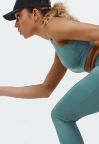 Woman in teal athletic wear and black cap leaning forward in a ready, athletic stance against a white background.