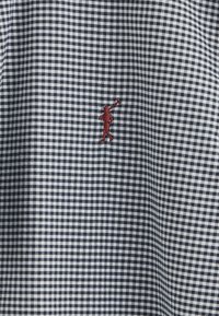 Marinblå och vit ginghamtyg med en liten röd broderad figur i en dynamisk pose, med en mjuk textur och tätt mönster.