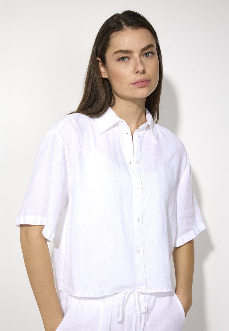 Witte linnen shirt met korte mouwen, knoopsluiting aan de voorkant, iets losse pasvorm en een afgeronde zoom. Heeft een opvouwbare kraag en een subtiele textuur.