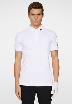 Witte polo shirt met een knoopsluiting bij de kraag, korte mouwen en een logo op de linkerkraag. Vergezeld van een grijze golfhandschoen op de linkerhand.