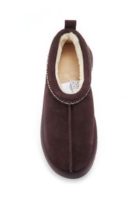 Hnědé nubukové pantofle s měkkou kožešinovou podšívkou, elastickým vrchem a dekoračním prošíváním kolem obvodu. Plochá podrážka vhodná pro použití uvnitř.