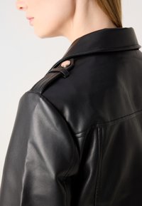 Veste en cuir noir avec un détail de bouton sur l'épaule latérale et une finition lisse et texturée. Le design comprend un col pointu et une couture au dos.